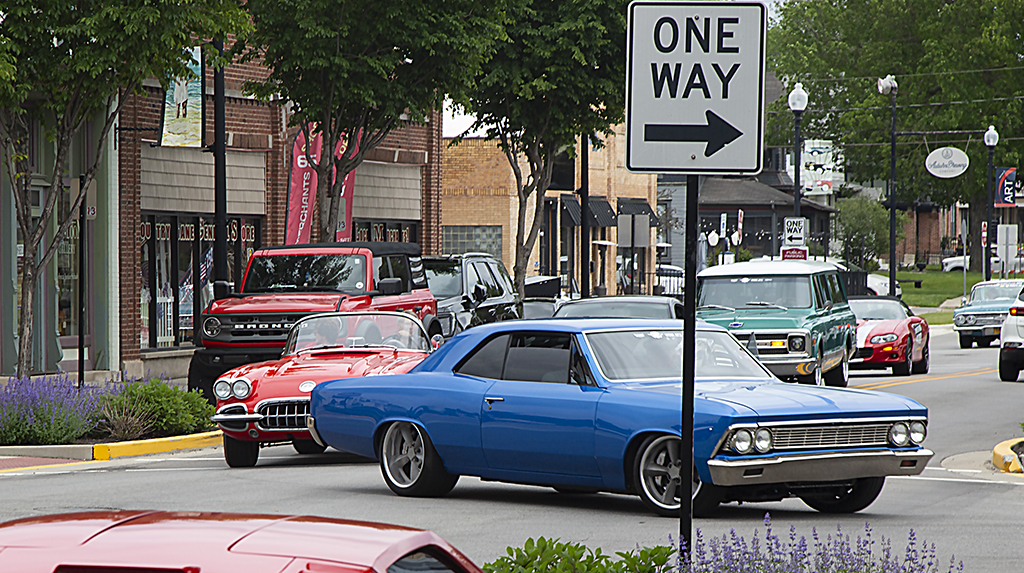 A 1966 Chevrolet Chevelle leads a line of classic cars and trucks driving slowly through a small downtown area